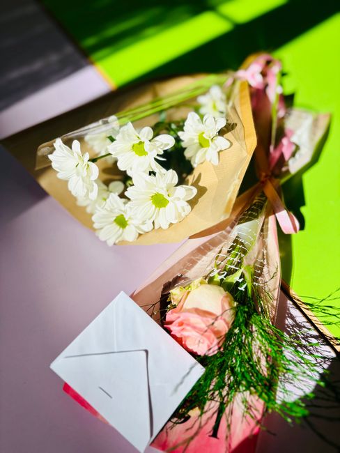 A floral arrangement featuring a bouquet of white daisies with yellow centers, wrapped in light brown paper and tied with a pink ribbon, complemented by a single pale pink rose wrapped in clear cellop