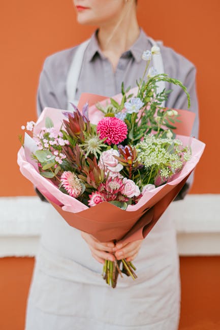 A person wearing a light grey shirt and a white apron holds a vibrant bouquet of fresh flowers, arranged as a floral gift for a birthday occasion. The bouquet features a variety of pink and white bloo