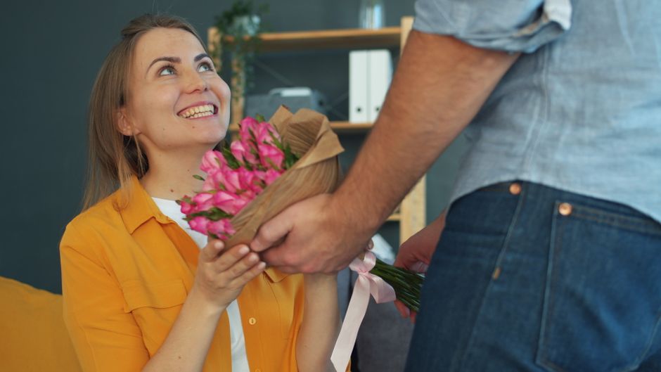 A woman with long blonde hair, wearing a mustard yellow shirt, is smiling and looking up while receiving a bouquet of vibrant pink flowers wrapped in brown kraft paper with a light pink ribbon from a 