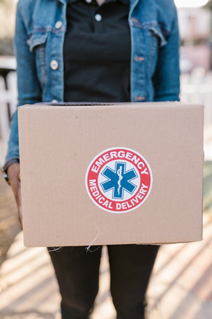 A person wearing a denim jacket and black top holding a cardboard box decorated with an 'Emergency Medical Delivery' sticker featuring a blue medical symbol and red border, set outdoors with blurred b