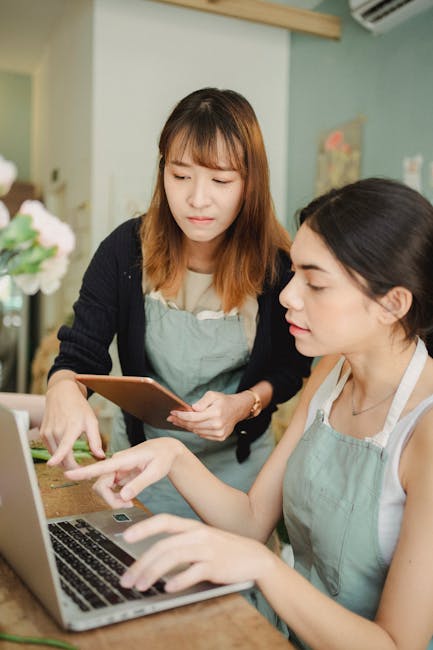 This image features two women in a florist shop or floral studio environment, engaging in a discussion about flower arrangements. One woman with shoulder-length light brown hair and wearing a light bl