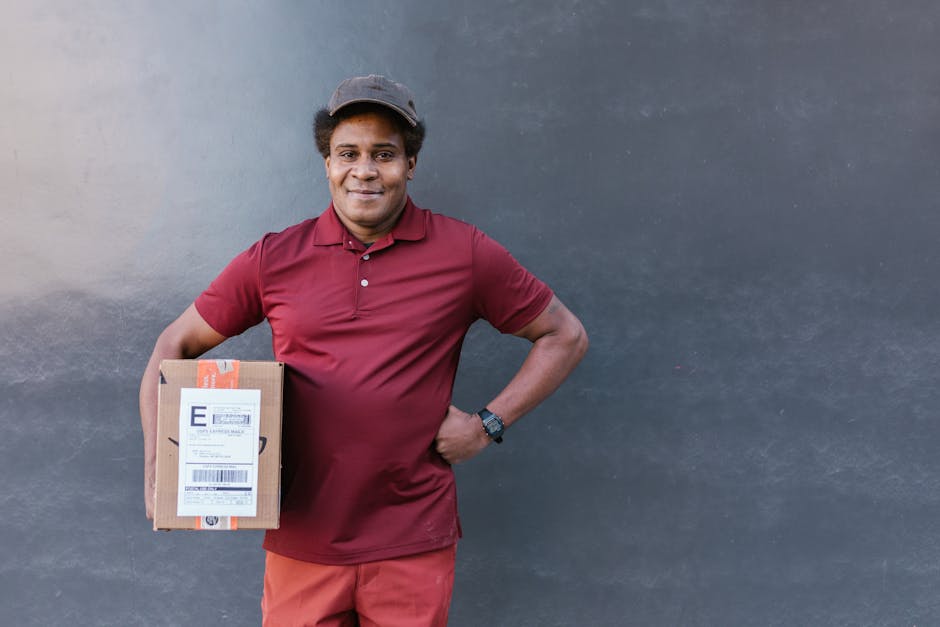 A male delivery courier stands against a plain grey wall, holding a small cardboard parcel with a shipping label in his left hand. He is dressed in a maroon polo shirt, red trousers, and a grey cap, s