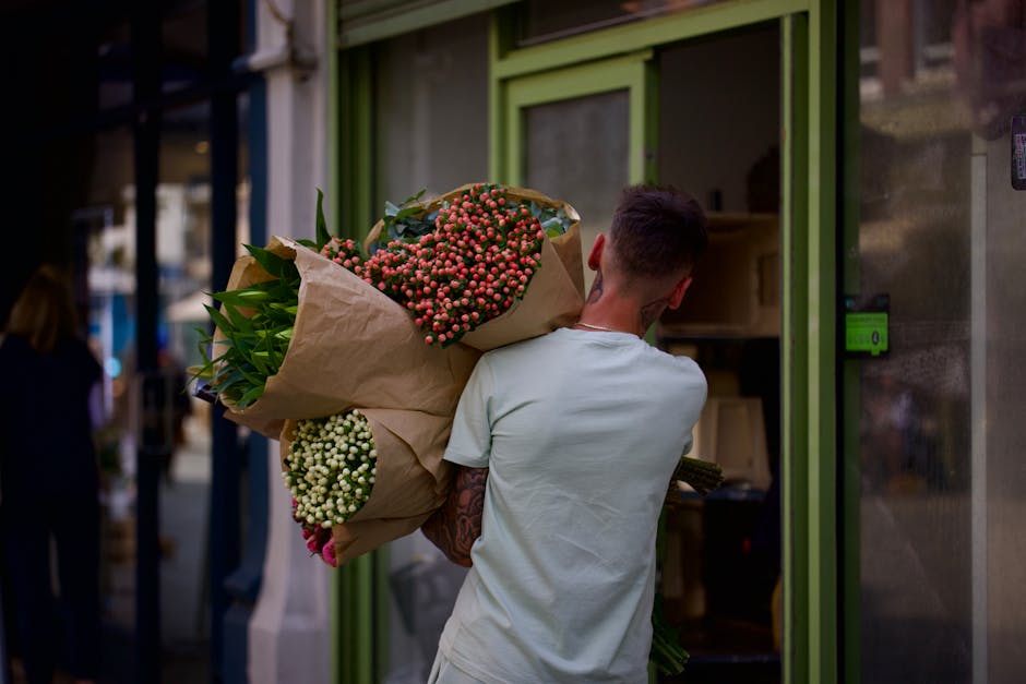 A man with short dark hair, wearing a light-colored t-shirt, is carrying a large bouquet of fresh flowers wrapped in brown kraft paper. The bouquet features clusters of small, round pink and white flo